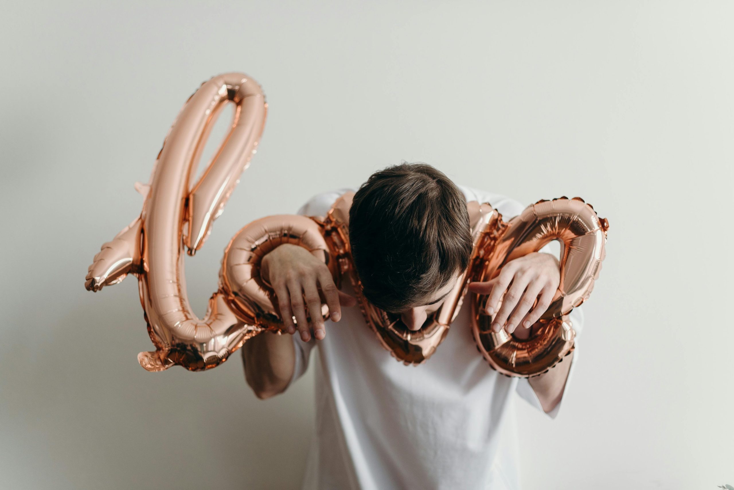 Un jeune homme en chemise blanche tenant des ballons « love » rose doré sur un fond neutre.