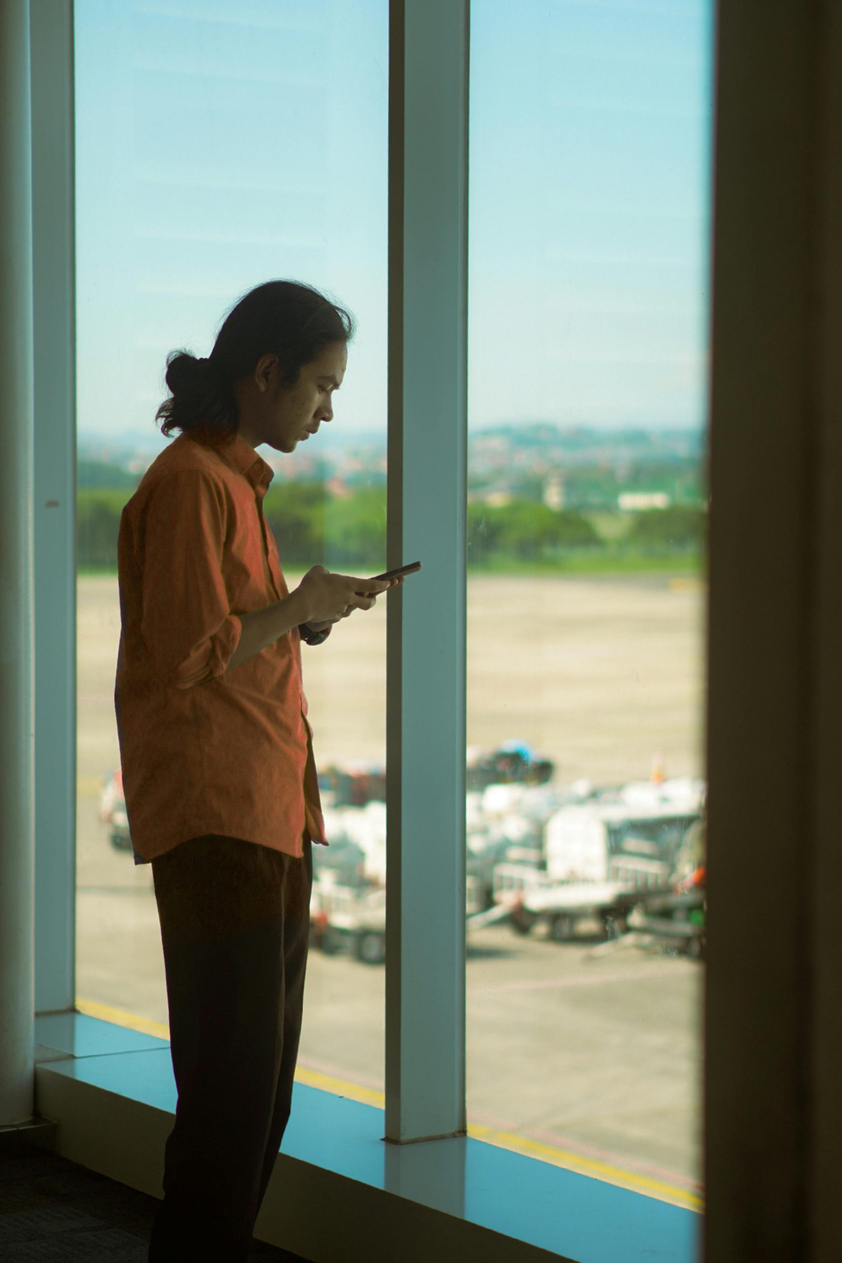 Homme en chemise orange utilisant un smartphone tout en se tenant près d'une fenêtre d'aéroport, avec vue sur la piste.