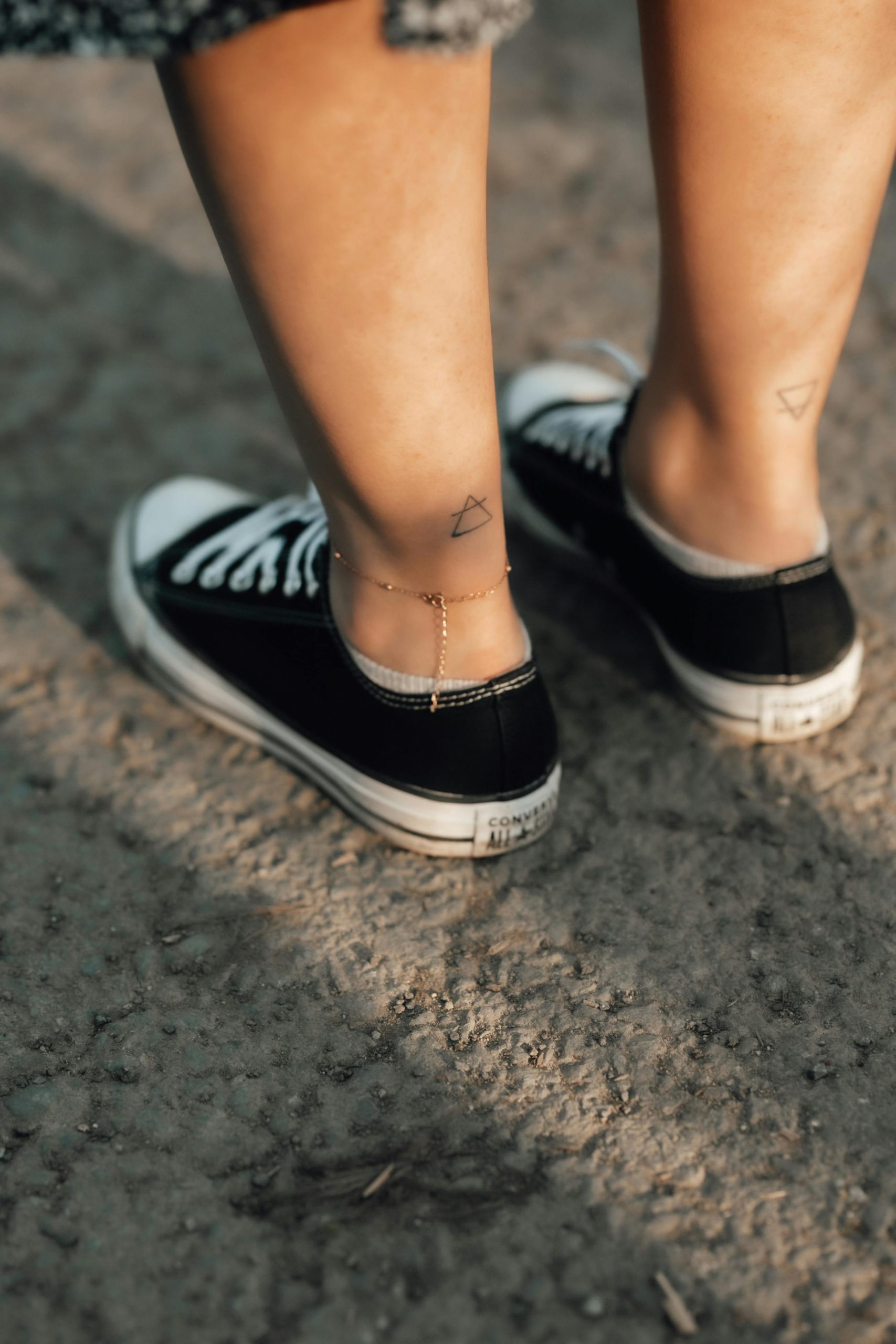 Close-up des jambes d'une femme, portant des baskets noires avec des bracelets de cheville et des tatouages, en extérieur.